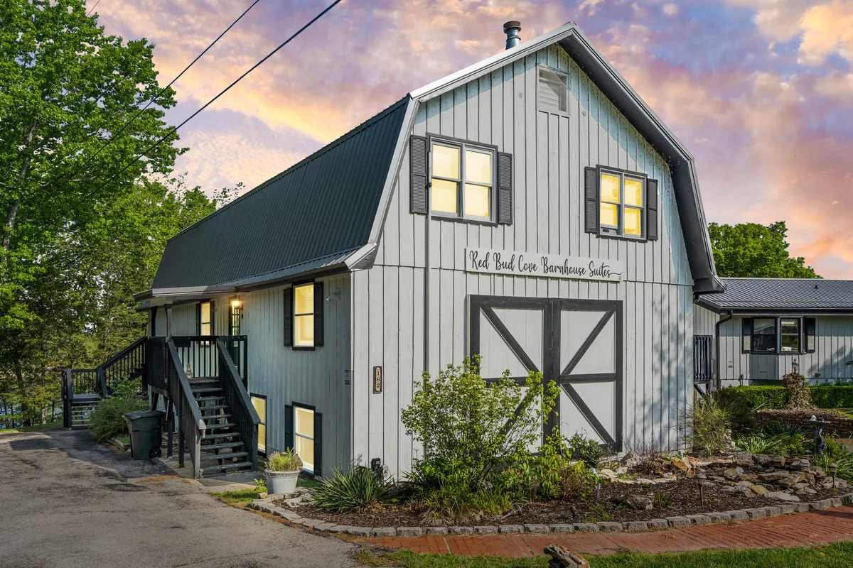 The hero image showcases the charming barn-style exterior of the Red Bud Cove Barn Suites Lodge at sunset. The building's warm, illuminated windows create an inviting glow, set against a beautifully landscaped garden. The scenic lakeside setting hints at tranquil evenings perfect for relaxing, stargazing, or socializing outdoors. The overall scene exudes a welcoming and peaceful atmosphere, making it an ideal spot for a memorable getaway.