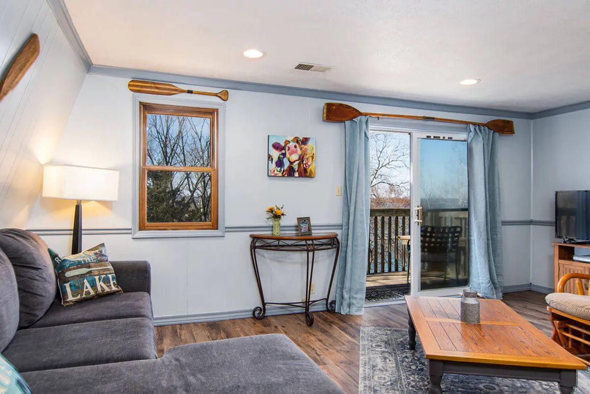 A cozy living room featuring a comfortable sofa adorned with a lake-themed cushion, wooden paddles as wall decor, a central coffee table, and, most notably, a sliding glass door leading to a deck with partial lake views. Natural light fills the space, inviting relaxation and offering a glimpse of the outdoors.