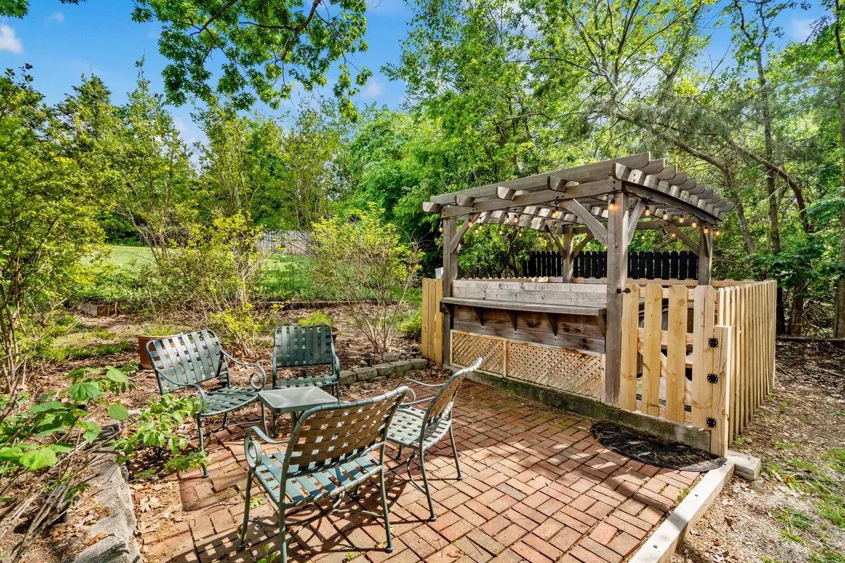 The image showcases a vibrant outdoor bar and seating area surrounded by lush greenery. The space is set up for social gatherings, with tables and chairs arranged for group interaction, perfect for outdoor dining, barbecues, or evening get-togethers. The overall atmosphere is relaxed and inviting, encouraging guests to come together outside.