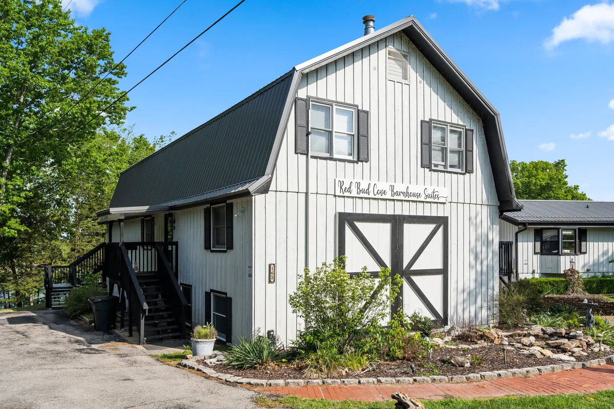 Exterior view of the Red Bud Cove Barnhouse Suite with barn-style architecture, visible signage, and a landscaped garden. The inviting porch and garden setting suggest a welcoming home base for exploration and gatherings.