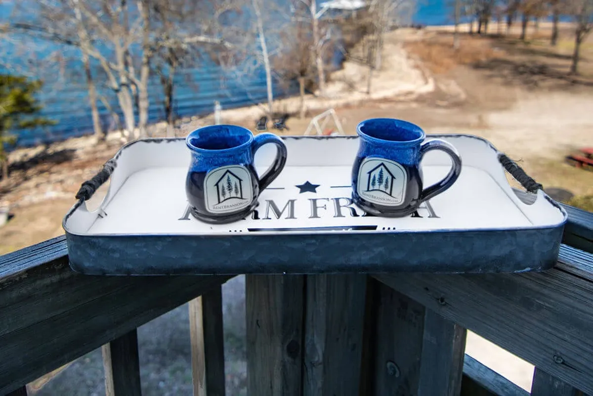 A cozy outdoor deck scene featuring a tray with mugs, a water view, and surrounding trees. The image is intimate, suggesting a morning coffee enjoyed together while overlooking the calm lake.