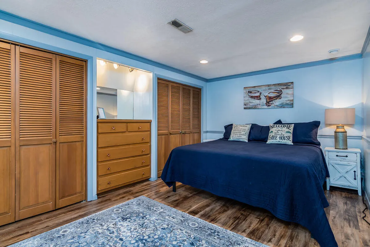 A cozy bedroom featuring a king-sized bed with plush bedding, a warmly lit lamp on a bedside table, a dresser, and nostalgic artwork on the wall. The soft lighting and carefully chosen décor create an inviting, restful atmosphere.