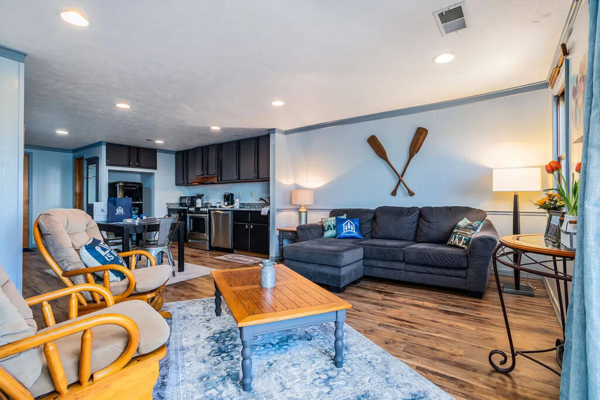 A warm, inviting open-plan living and dining area with a sofa, wooden coffee table, visible kitchen appliances, and decorative paddles on the wall. The space is designed for relaxation and socialization, with plenty of natural light and cozy furnishings.