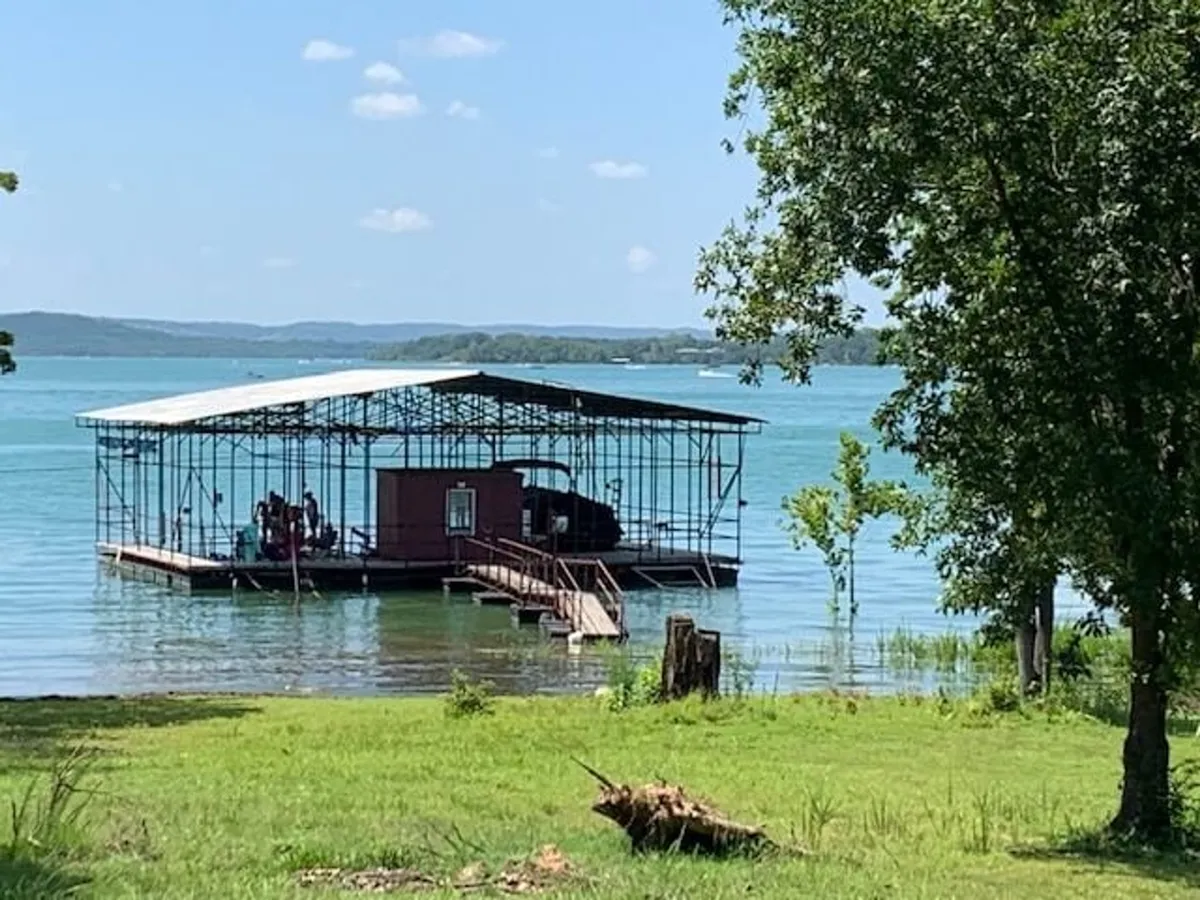 A relaxed scene featuring a floating dock, a boathouse, tranquil lake waters, and natural greenery. The setting invites images of swimming, fishing, or simply enjoying the view with others.