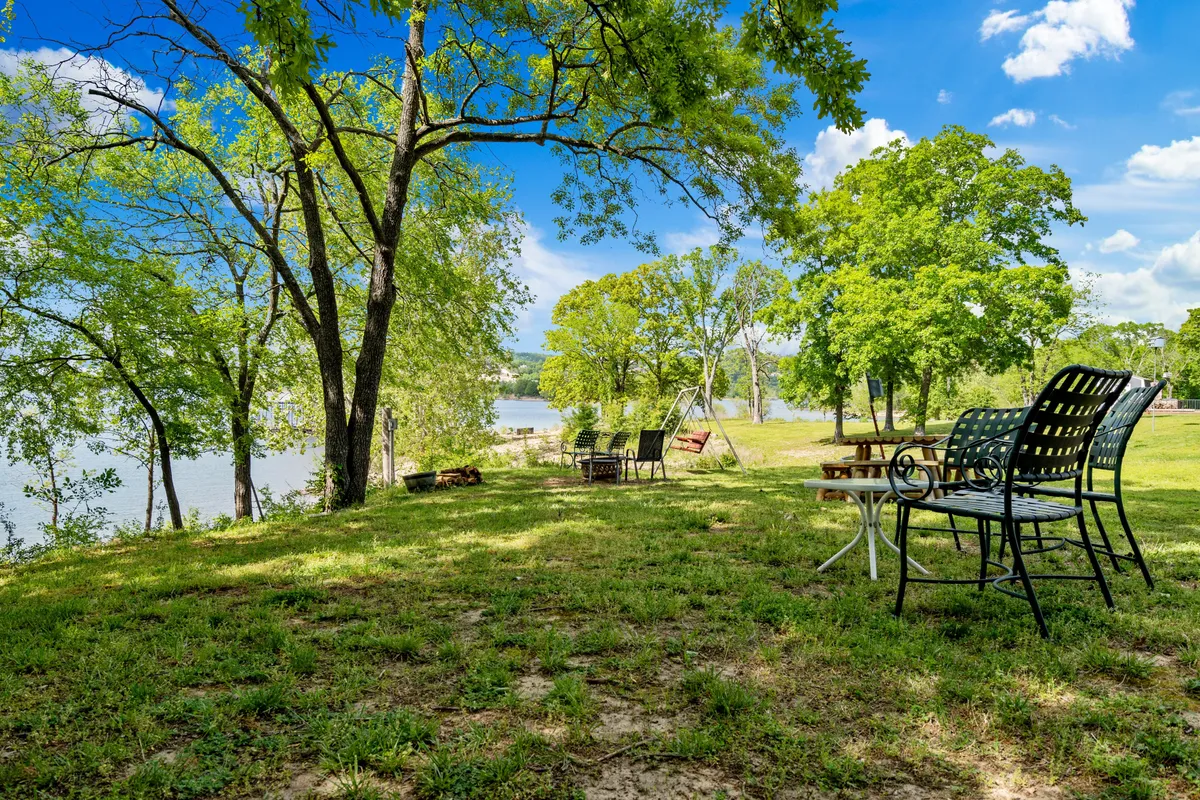 This image features a peaceful riverside lawn with green grass, tall trees, a swing set, and lawn chairs overlooking a river. The space feels welcoming and playful, suggesting an ideal spot for children to play and families to relax together near the water.