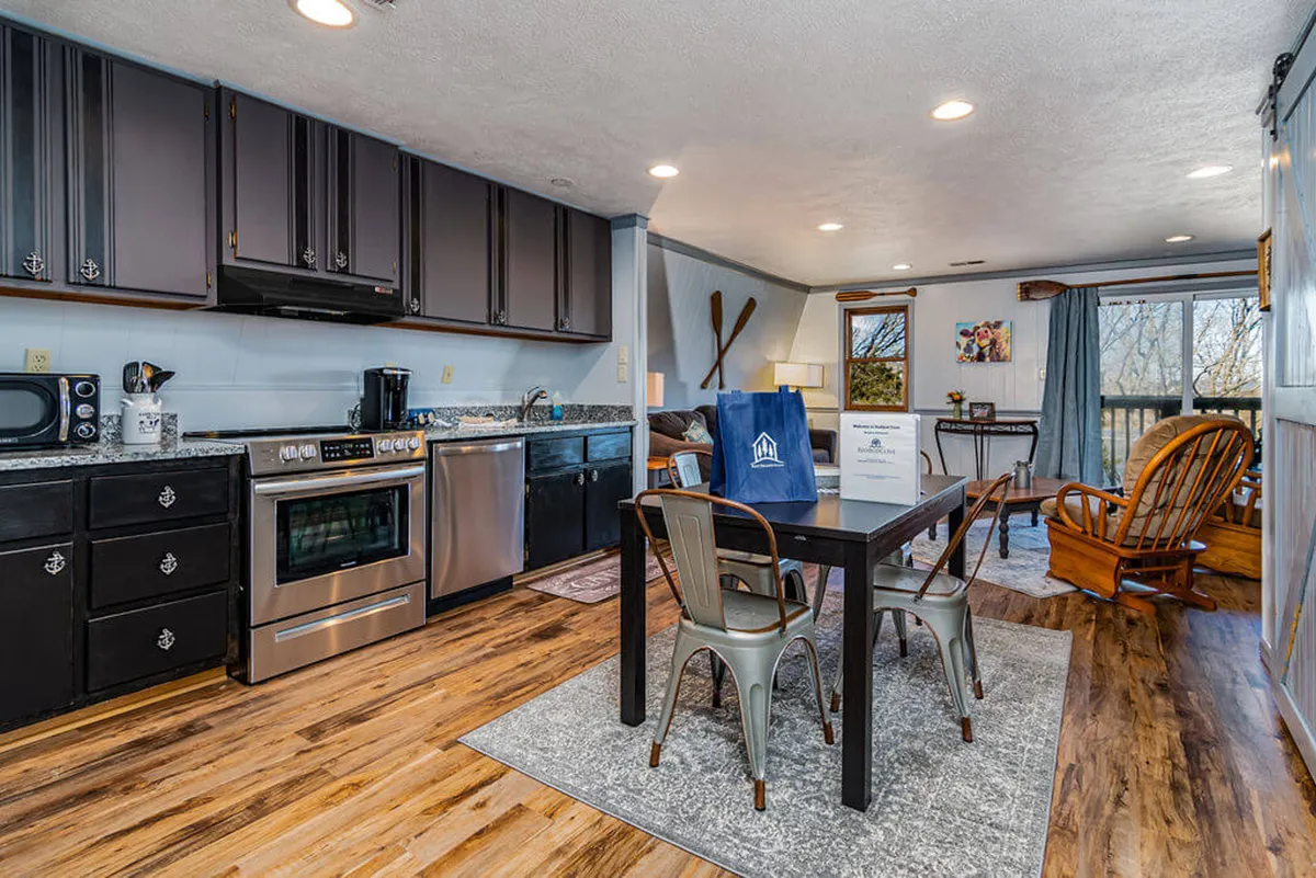 This inviting image captures a cozy interior kitchen and dining area. The space features kitchen appliances and a dining table set near large windows, with a rocking chair nestled nearby. The overall feel is warm and welcoming, with natural light streaming in and a layout that encourages socializing and togetherness.