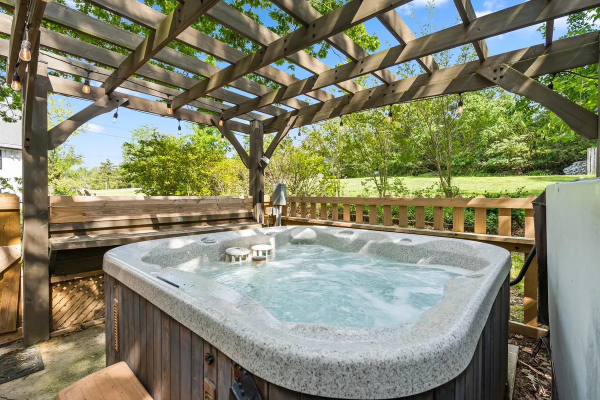 This image showcases a stylish outdoor hot tub beneath a pergola, surrounded by lush greenery. The setup hints at opportunities for relaxation, stargazing, and socializing with fellow guests, with enough space nearby for barbecue activities.