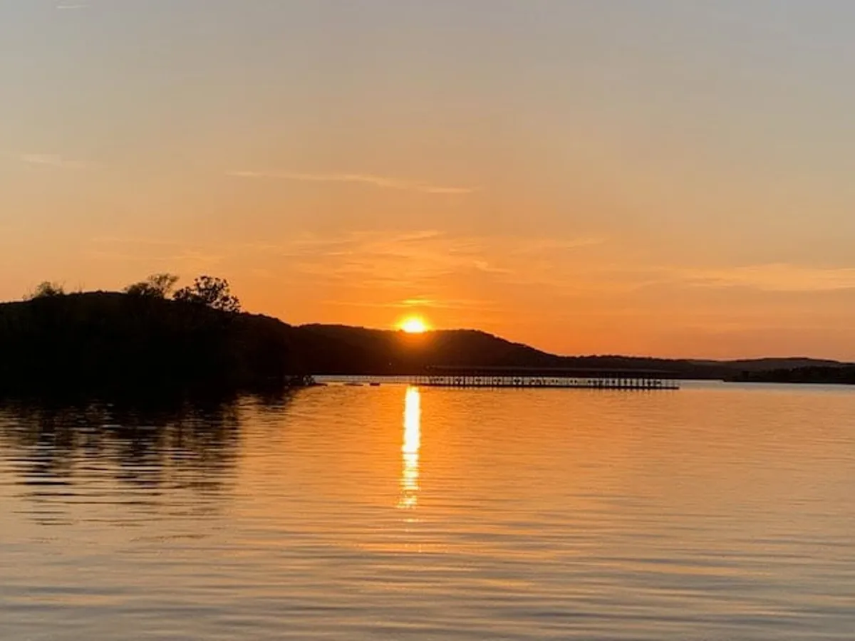 This image captures a serene lakeside sunset with the sun reflecting off calm water, a distant horizon, and a picturesque pier. The tranquil mood and warm colors suggest a peaceful, romantic setting ideal for couples' relaxation and shared moments in nature.