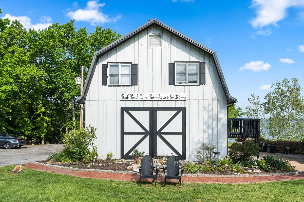 This exterior shot highlights the welcoming barn-style building of Red Bud Cove Barnhouse Suites, with its distinctive sign and two inviting chairs set out front. The image conveys a sense of relaxation and socializing, providing a clear sense of arrival and community.