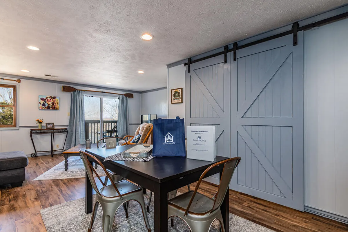 A cozy dining table stands in the foreground, illuminated by natural light from a window with a view. Sliding barn doors add rustic character, and the overall mood is one of intimacy and warmth—ideal for family meals, game nights, and heartfelt conversations.